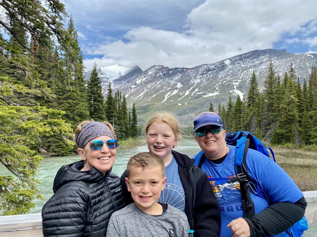 Hillary Hatcher and her family, two kids and another adult, smile and pose on a bridge in front of a river and mountains with a dusting of snow