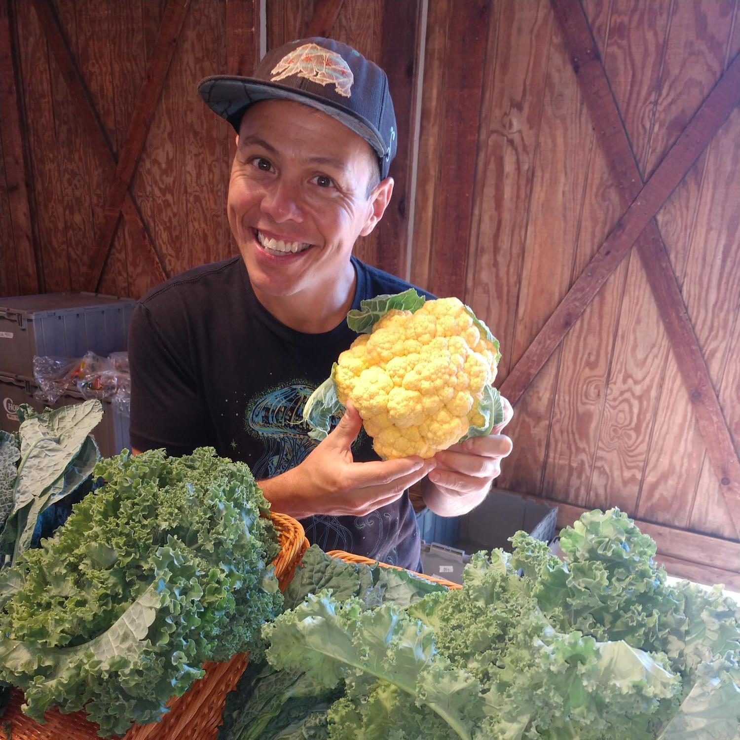 JJ Neville holding up a cheddar cauliflower. There are other greens on a table in front of him