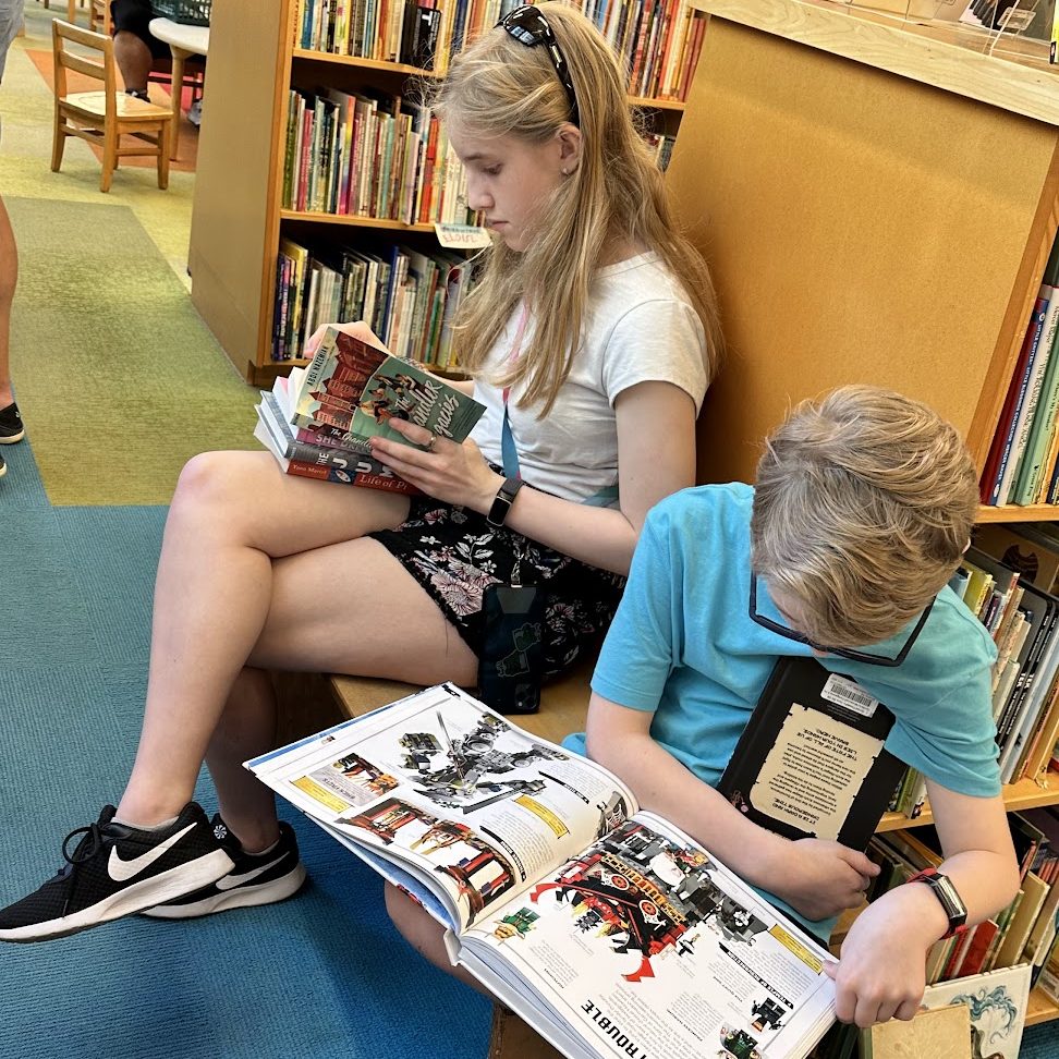 a boy and girl sitting on a wood bench inside a bookstore, they are both reading a book and have others in their lap.