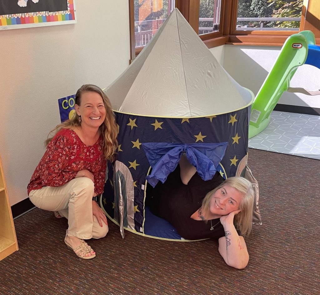 Trish Byrne and Courtney Yeagers smile for the camera. One is sitting outside a child-sized play tent, and the other is lying down inside with her head and shoulders sticking out.