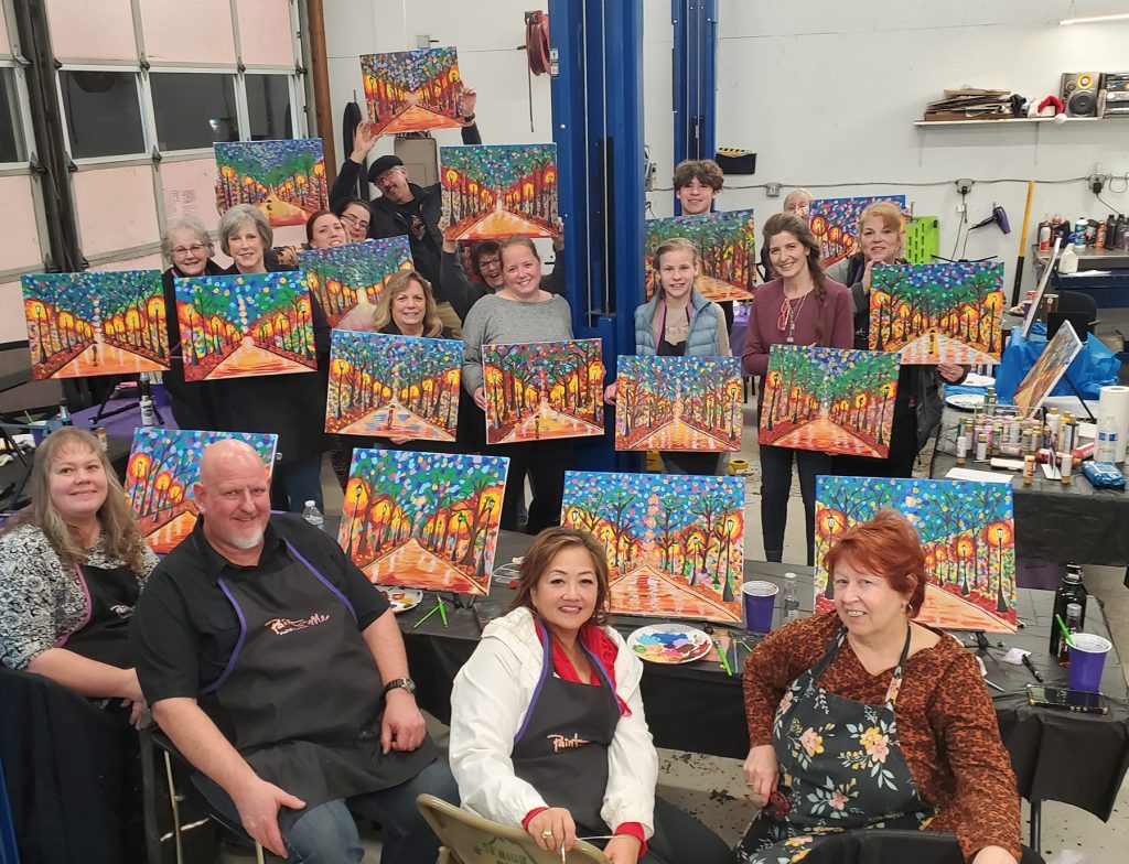 people sitting and standing by tables with their paintings - all the same of a path with trees and a sky - and smiling for the camera