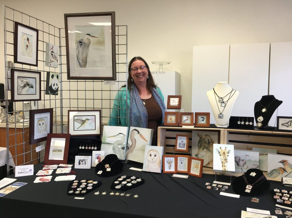 Nancy Broach stands behind her table full of her art and jewelry and smiles for the camera