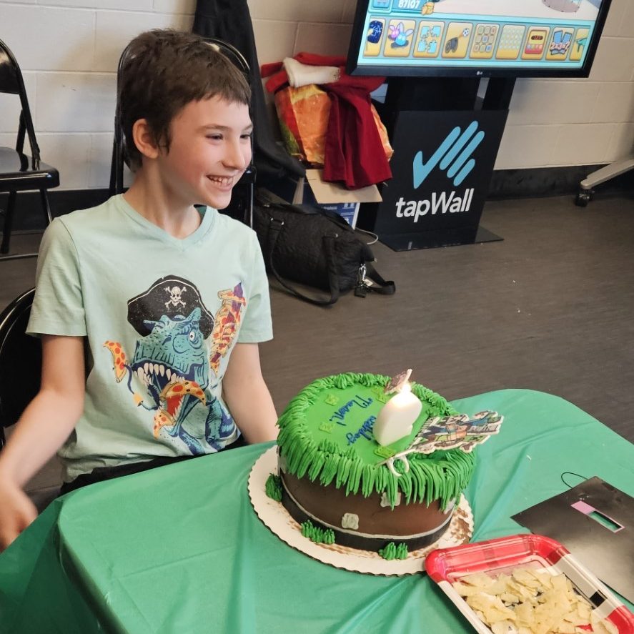 A kid sitting at a table with a dino shirt one, smiling at his birthday cake that is brown and green