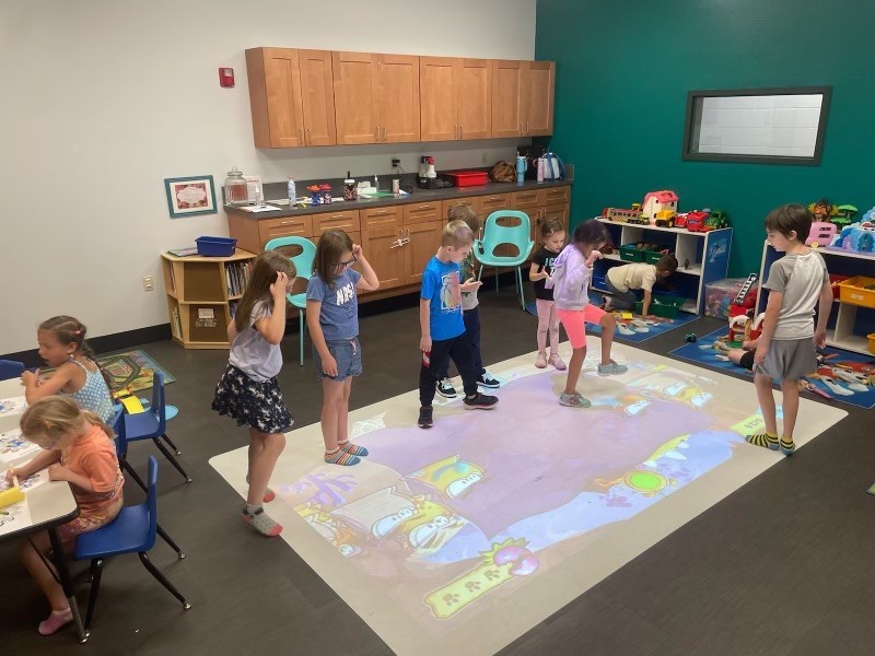 kids in a room with a cartoon projected onto the floor. Some kids are sitting at tables coloring