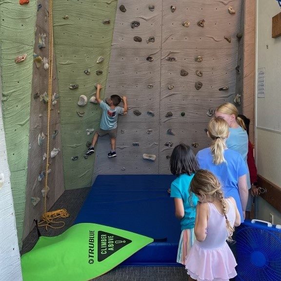 A kid on an indoor rock climbing wall with kids on the ground in a line