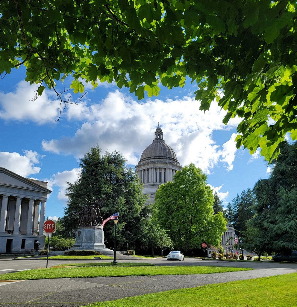 the Olympia Capitol building behind some green trees with an american flag and a statue of soldiers out front