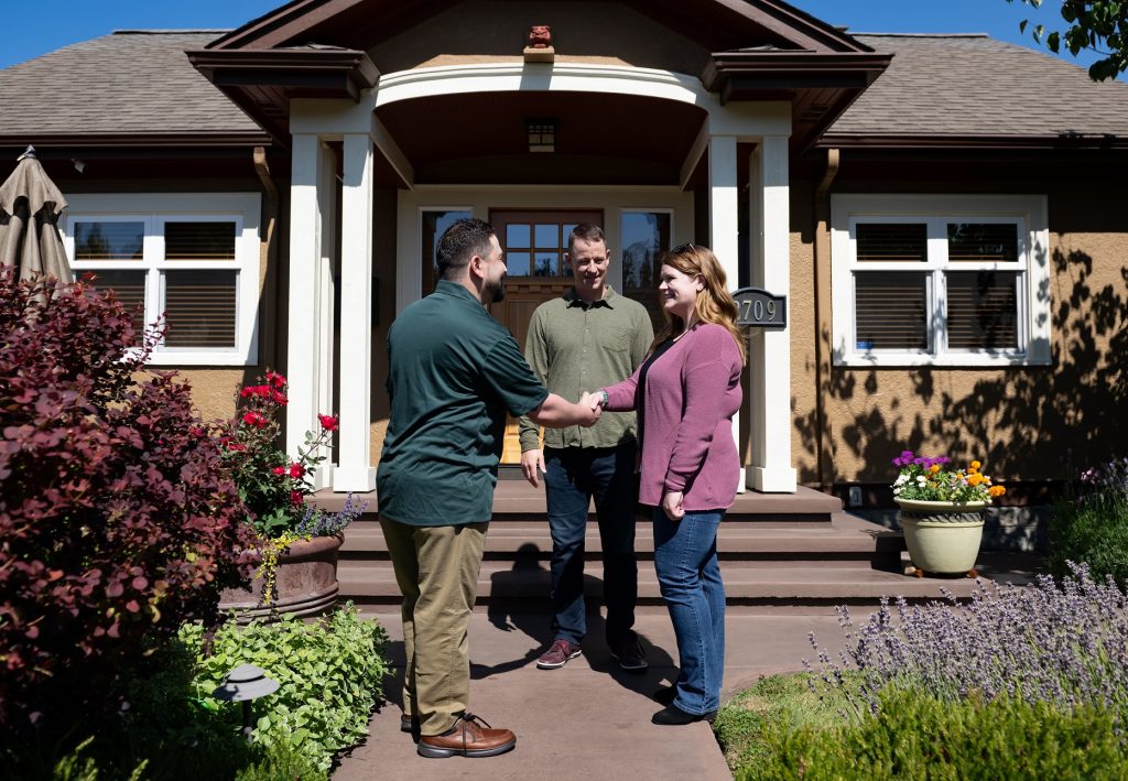 A man shakes hands with a woman, who is standing by another man outside a house.