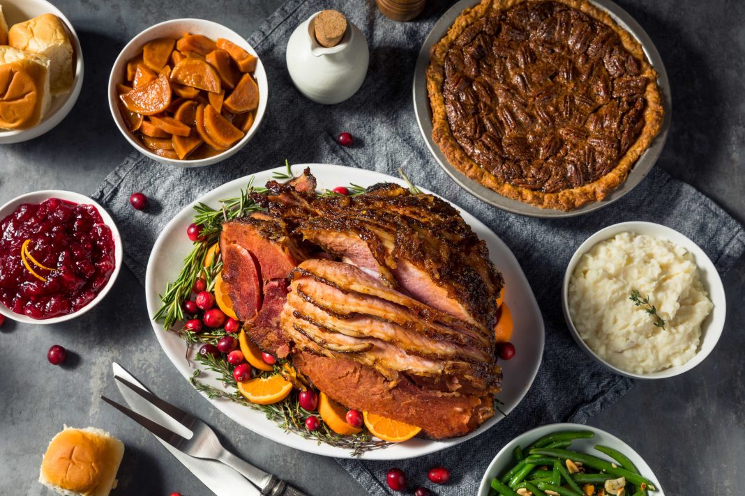 a table with plates full of traditional holiday dinner food items, like ham slices and a pecan pie