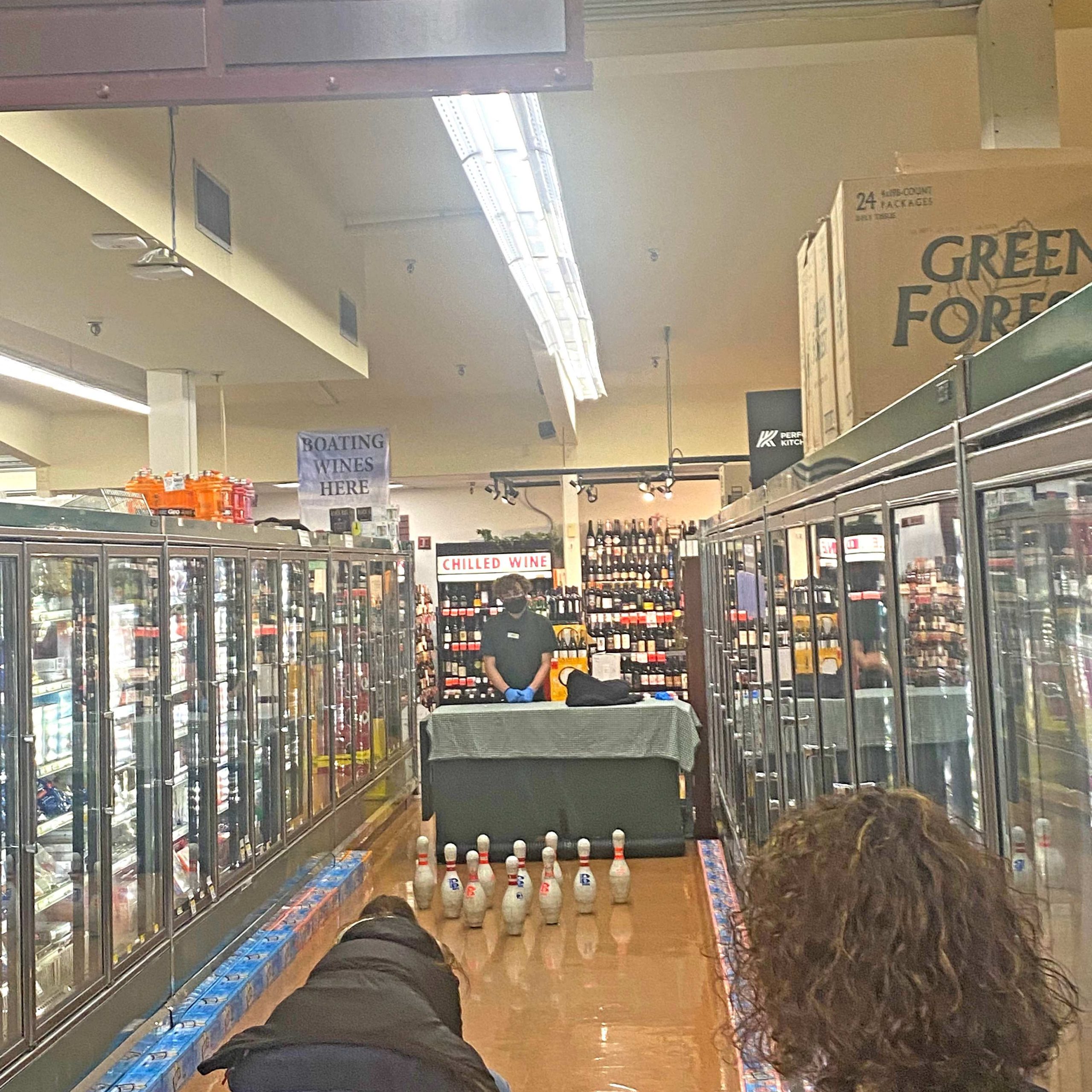 Freezer aisle of Thriftway set up with a table and a coworker and 10 bowling pins with a customer seen bent over about to bowl.