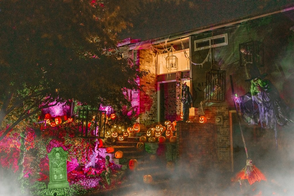 a house decorated for Halloween with pumpkins, skeletons, witches and purple and green lights, with fog over it.