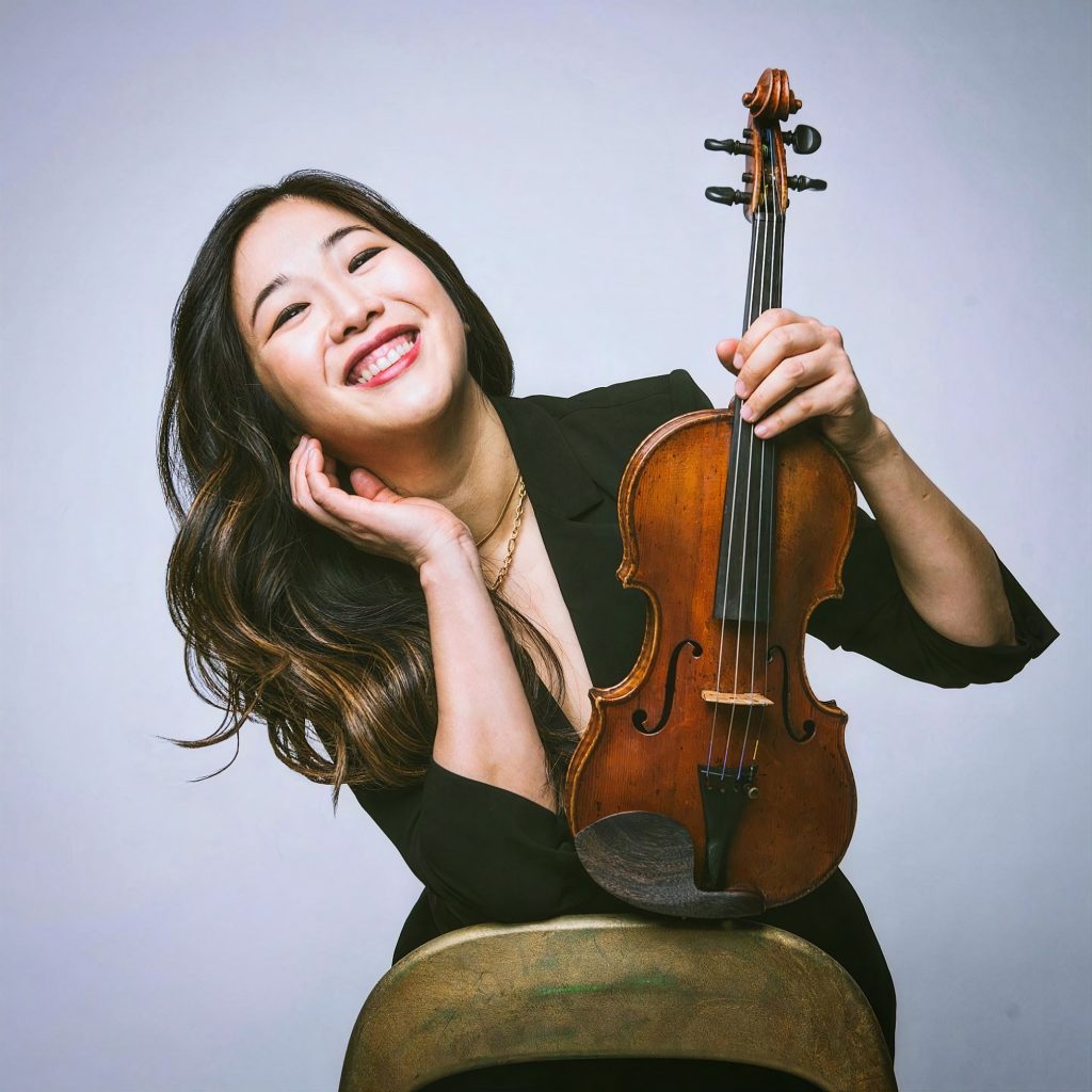 Kristin Lee leaning against a chair holding her violin