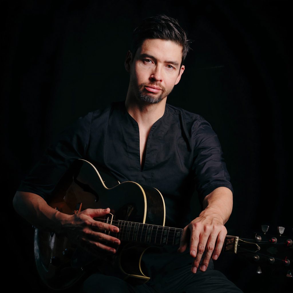 Miles Okasaki sitting in a chair holding his guitar against a black background