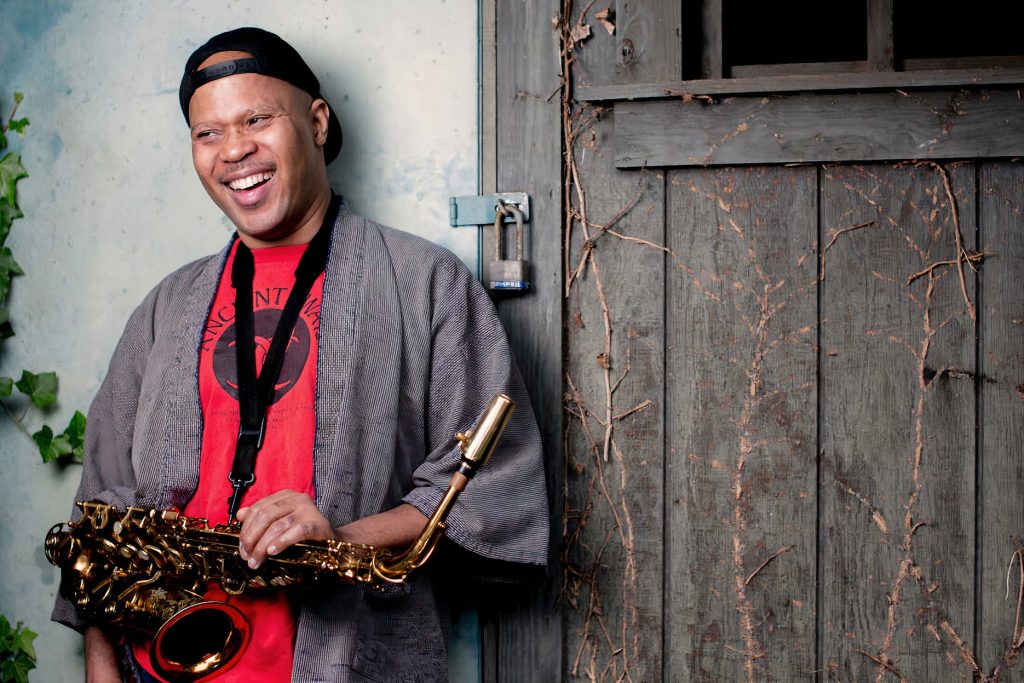Steve Coleman leaning against a barn door laughing with a sax around his neck