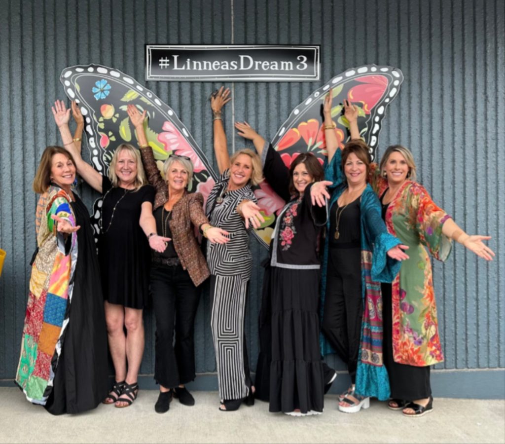Seven woman posing with their arms outstretched in front of a large butterfly painting on a wall with the words, '#LinneasDream3' on it