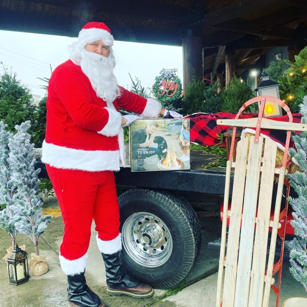 Santa by a truck with presents, a sled and Christmas trees around him