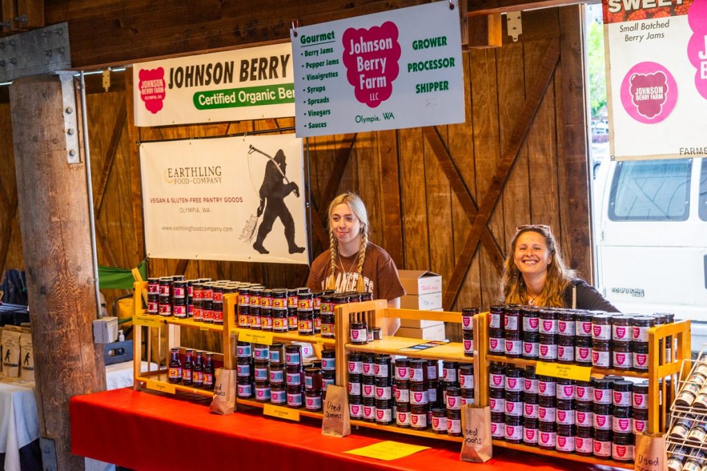 two women sitting at the Johnson Berry Farm booth at the Olympia Farmers Market with jars of jams and jellies.