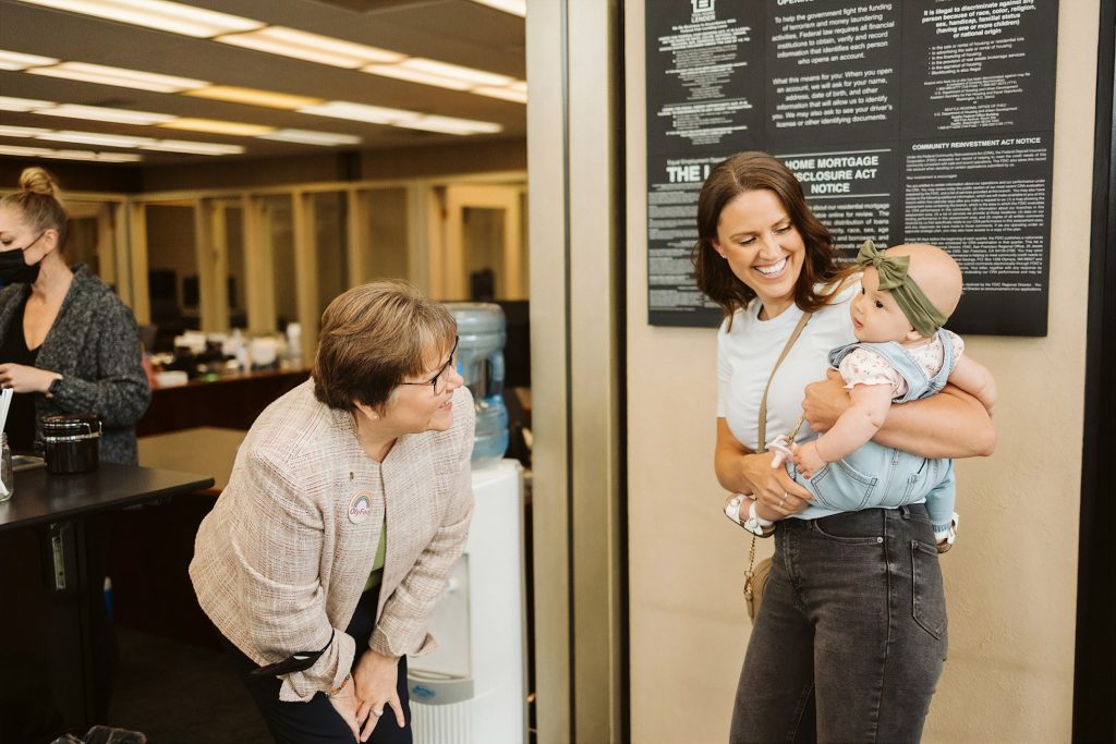 a woman bends down and smiles at a baby that a woman is holding on her hip