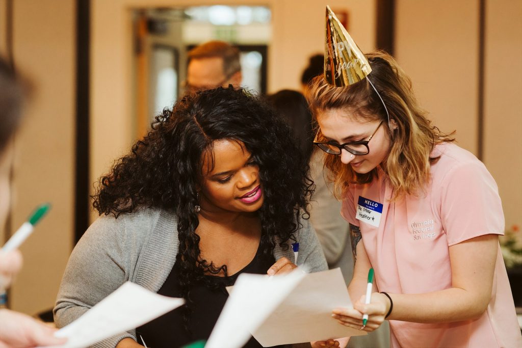 two OlyFed employees look at a paper together, one is wearing a party hat and a name tag