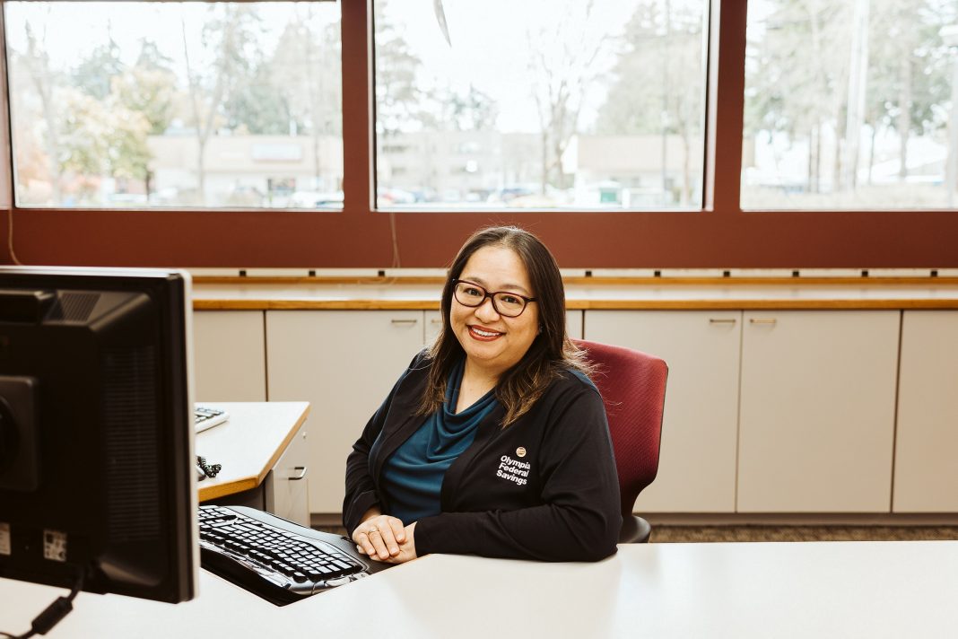 a woman smiles while sitting at a desk, she is wearing an OlyFed sweater
