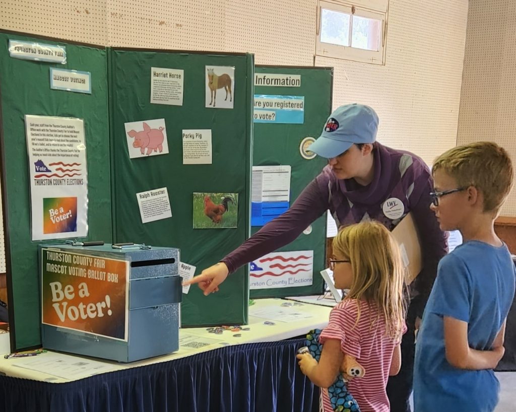 A woman shows a boy a and girl a ballot box at a booth with signage and information about voting