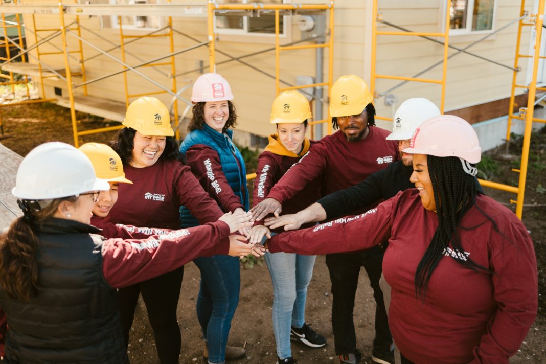 a group of habitat for humanity workers in matching sweatshirts and hard hats, standing in circle with one hand stretched into the center, stacked on top of each other.