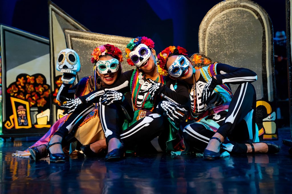 Three people in sugar skull costumes sit on a stage, one holds up a fake skull.