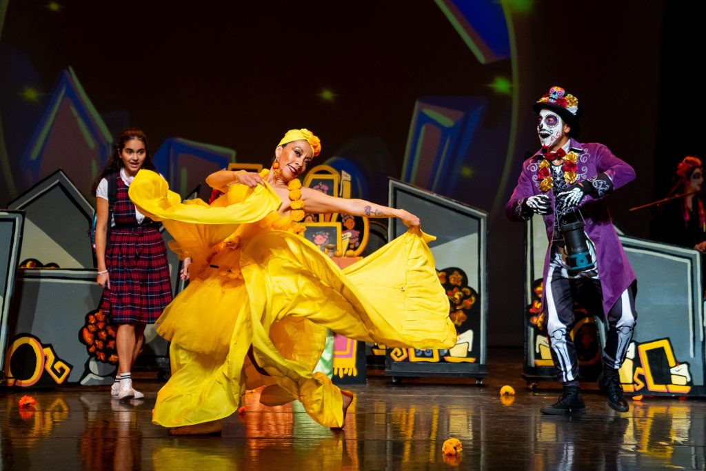 A woman in a full yellow dress dances on stage with a girl in a plaid uniform and a man in a sugar skull outfit standing nearby