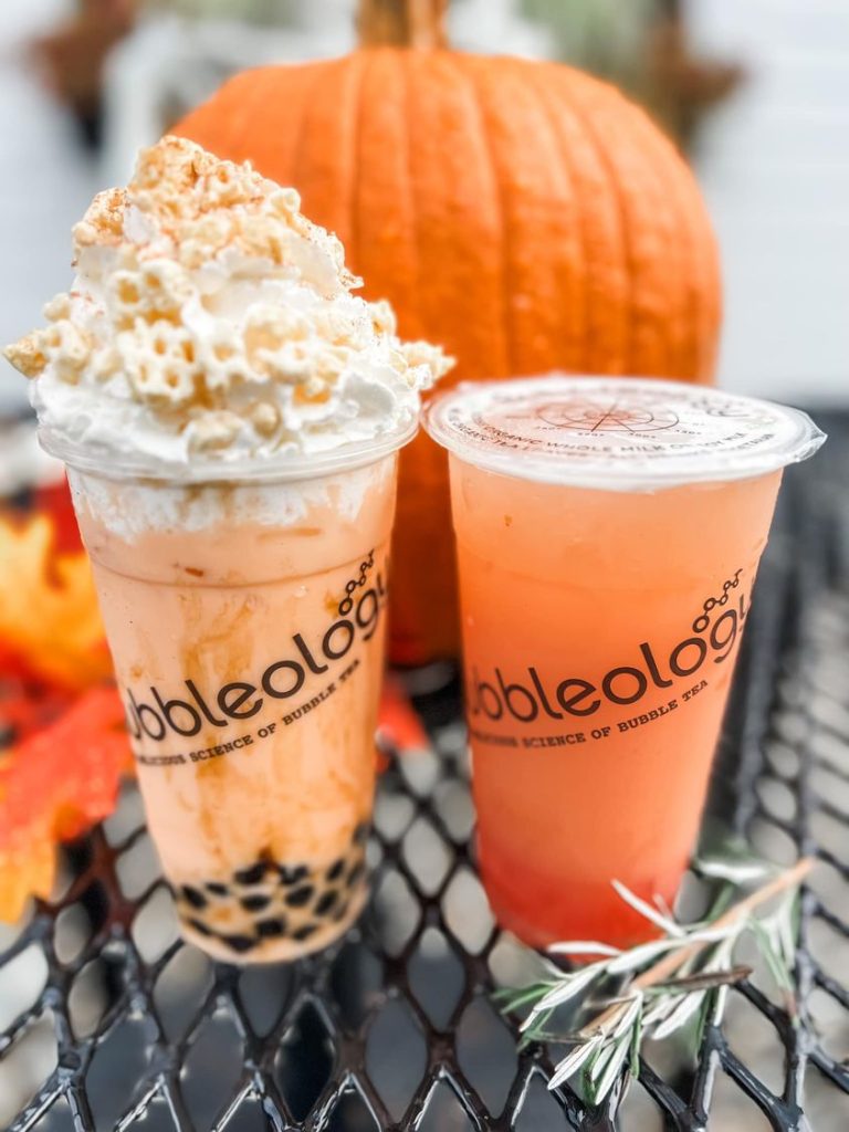 2 bubble tea drinks, one with whipped cream, on a metal table with a pumpkin behind them
