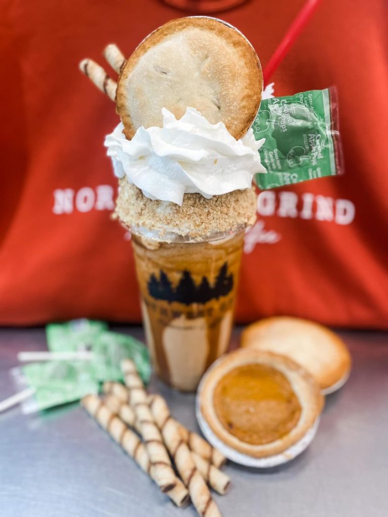 a 32-ounce blended with spiced chai drizzled with pumpkin sauce and topped with whipped cream, a caramel apple sucker, two rolled cookie wafers, and apple and pumpkin mini pie sitting next to it with a lid rolled in graham crackers