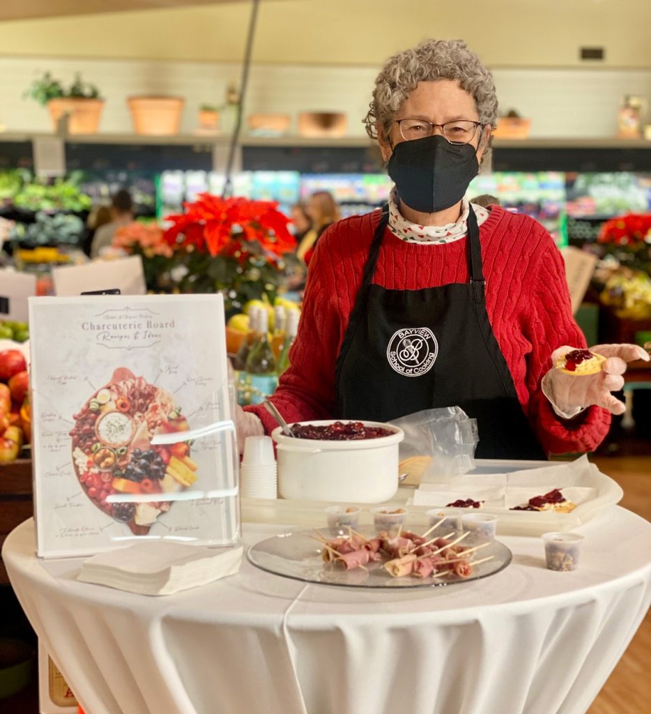 a woman in a mask and an apron stands at a high table with food samples on it