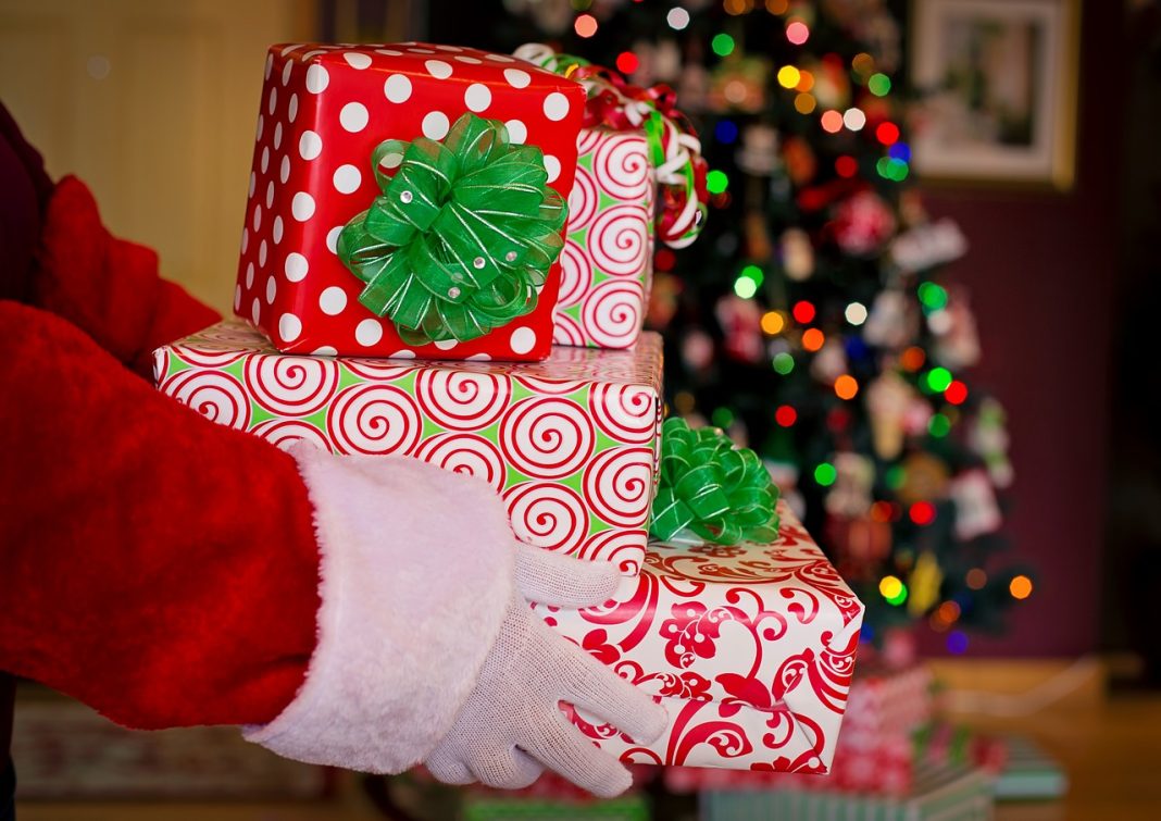 santa hands in white gloves holding three presents wrapped and red and white paper with a Christmas tree in the background