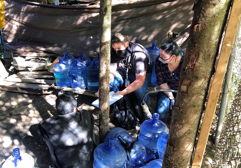 Policewoman kneeling a homeless camp with a person and lots of jugs of water around them.