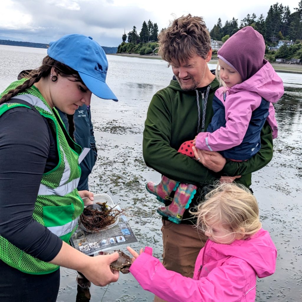 a man carrying a child and a woman and a child standing on the beach looking at a crab the woman is holding