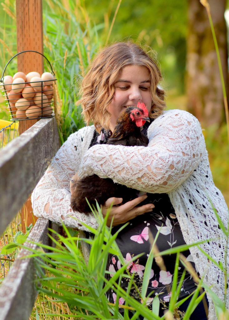 Holly Johnson in a field of tall grass by a wood fence with basket of eggs on it. She is holding a black chicken