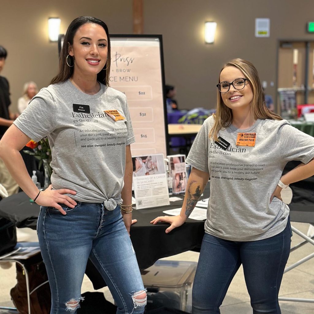 Two woman in shirts with the definition of Esthetician on them standing in front of a table at a career fair