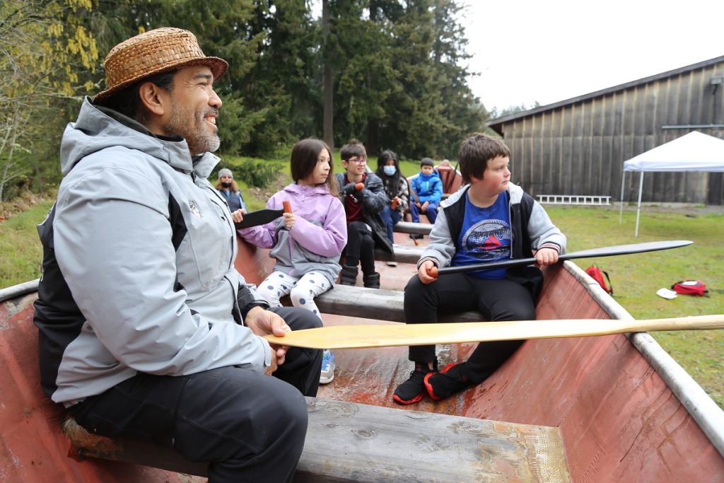 Kids are sitting in a canoe in grass with an adult holding an oar in front