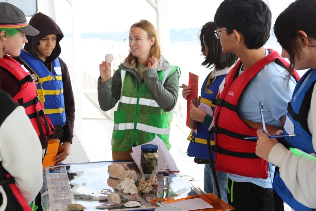 Kids with life vests stand in front of a table with a bunch of seashells and other specimens. One holds up a sand dollar