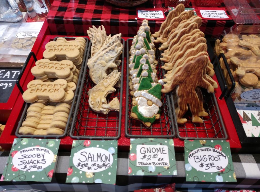 dog cookies in rows in metal racks. An assortment of shapes including a bigfoot, frosted gnome, a salmon and a bone that says 'scooby snack'