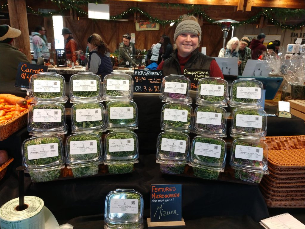 woman smiling behind a display rack full of plastic containers of microgreens