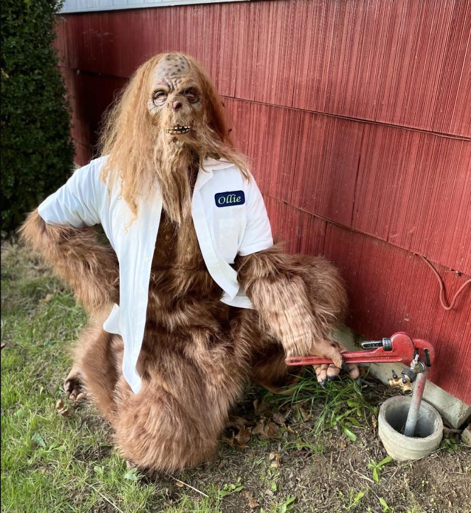 a person dressed as Bigfoot kneeling by a house with a wrench on a spigot