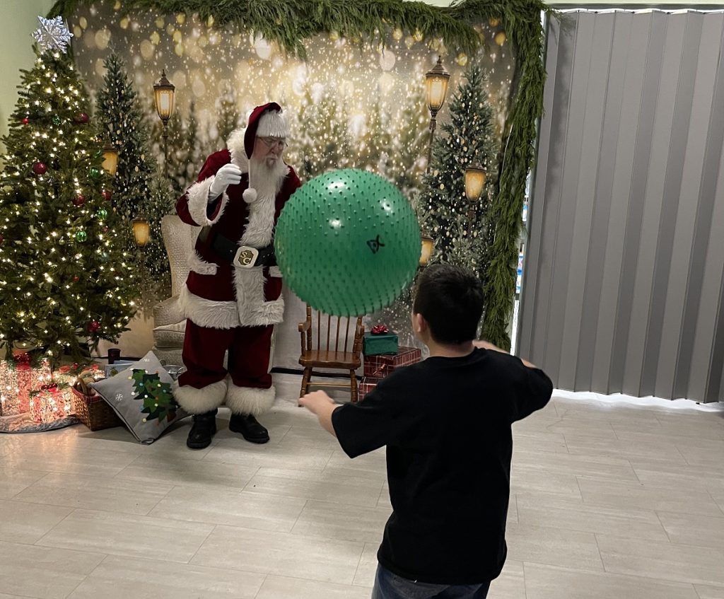 Santa playing catch with a big green ball and a young boy in a room with low-lit Christmas Tree