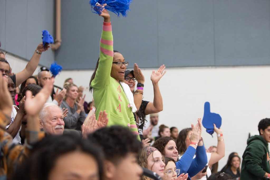 Fans cheering on the SPSCC team with blue pom poms and foam figures. One fan is standing up in the crowd