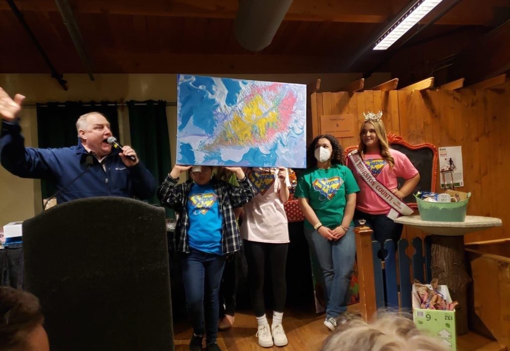 A man on a microphone stands next to people holding up a painting, a girl on the side is wearing a crown and a sash that say, 'Miss Thurston County.'