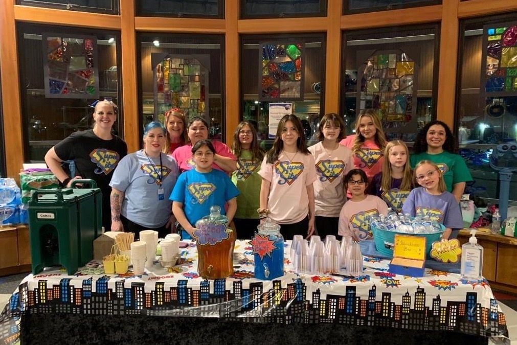 Large group of kids standing behind a table with different non-alcoholic drinks on it