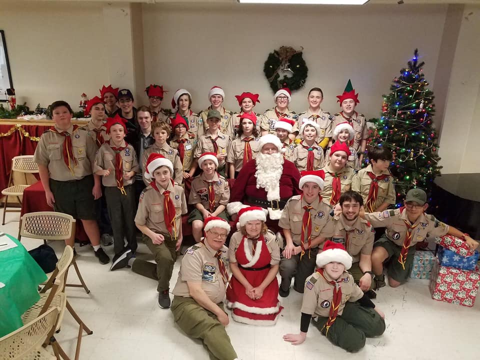 Boy Scout troop in uniforms gathered around Santa for a photo with a christmas tree and presents.