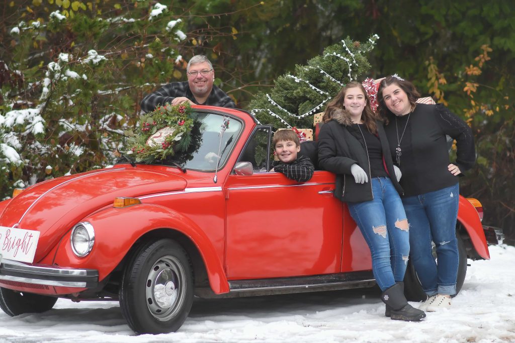 Holly Johnson and her family posing around a red vw bug convertible with a christmas wreath on it's hood and presents and a Christmas tree in the back