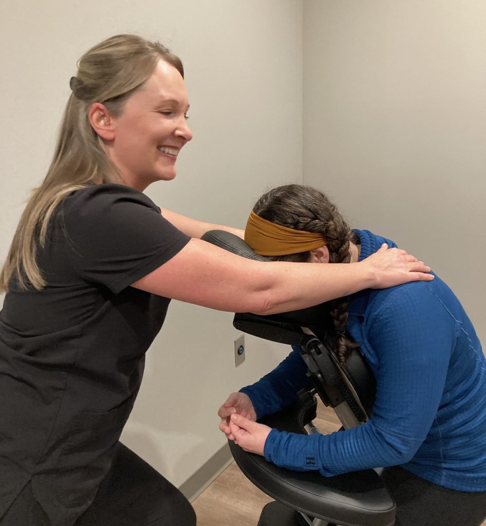 massage therapist standing in front of a person in a massage chair, with their hands on their shoulders