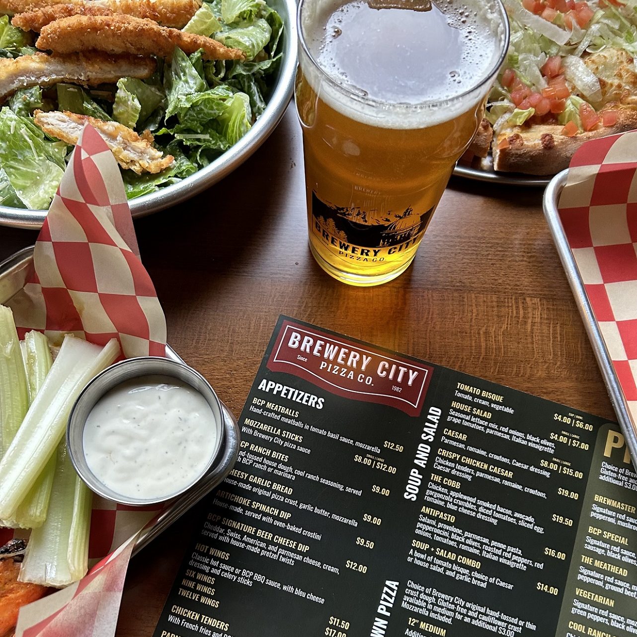 vegetable sticks with ranch dressing, chicken wings, beer and a menu on a table.