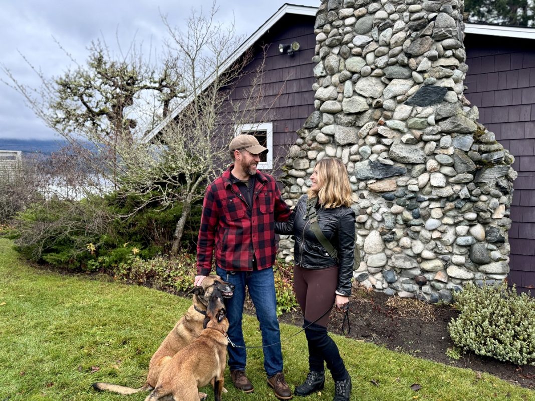 A couple stand in front of a rock and wood building with a dog by their side