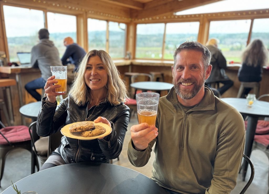 two people sit at a table in Finnriver Farm and Cidery. One holding a plate of food and the other a drink.
